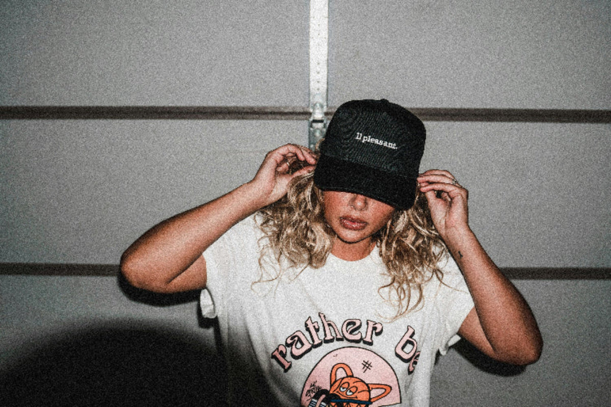 Person wearing a white t-shirt with graphics and text, holding a black cap.